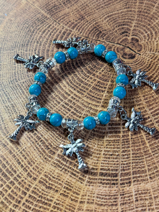 A beaded stretch bracelet with bright turquoise-blue beads, silver spacer beads, and multiple silver palm tree charms, displayed on a textured wood slice background.